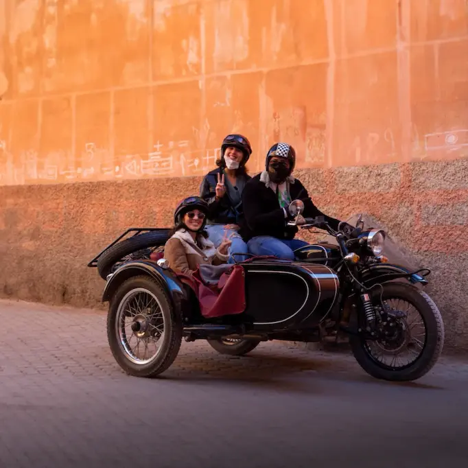 Three people riding a motorcycle in Morocco with a sidecar along a street with an orange wall.